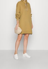 Olivgrön hoodie-klänning med en krage, långa ärmar och framzipperedetaljer. Matchad med vita sneakers och en beige väska.