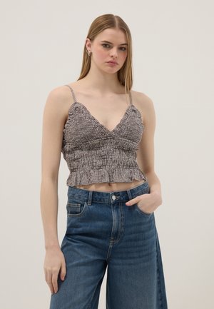 Jeune femme aux longs cheveux lisses portant un crop top smocké sans manches et un jean bleu large, debout avec une main dans la poche.