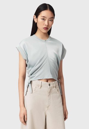 Młoda kobieta o długich, ciemnych włosach nosi jasnoniebieski krótki top z wiązaniami po bokach oraz beżowe spodnie z wysokim stanem na jednolitym tle.