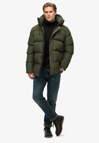 Olivengrøn pufferjakke med høj krave og hætte, med lynlåslukning og elastiske manchetter, båret over en sort turtleneck og blå jeans.