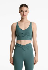 Teal színű, varrat nélküli sportmelltartó széles pántokkal és V-nyakkivágással, elől ráncolt díszítéssel, párosítva hozzá illő magas derekú leggingssel.