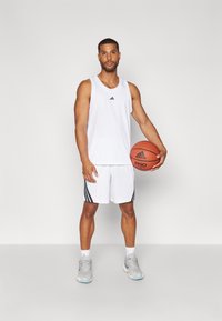 Homem vestido com um fato de basquetebol branco da Adidas, segurando uma bola de basquete, de pé contra um fundo claro e simples.
