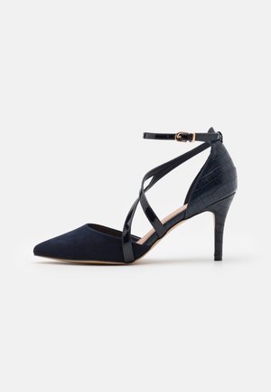 Chaussure à talon aigu en bleu marine, avec un bout pointu, en daim et en cuir verni. Présente des accents en relief crocodile et des brides croisées à la cheville.