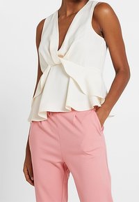Kermansävyinen peplum-paita, jossa syvä V-pääntie ja kerroksellinen röyhelömuotoilu, yhdistetty vaaleanpunaisiin housuihin, joissa on sileä kangas ja rento istuvuus.