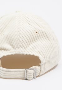 Gorra de pana crema con textura acanalada, correa ajustable en la parte posterior y detalles de hardware plateado. Diseño limpio y sencillo.