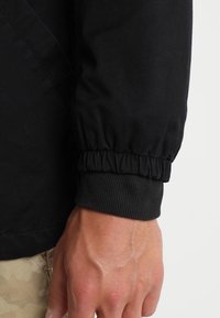 Czarny mankiet kurtki z elastyczną, zebrana taśmą, wykonany z gładkiego materiału o subtelnym połysku, prezentujący zbliżenie tekstury tkaniny.