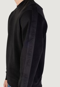 Hombre vestido con una sudadera negra con cinta del logo negro de Armani Exchange corriendo a lo largo de la manga exterior, de pie contra un fondo neutro.