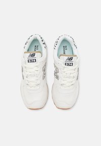 Paire de baskets blanches New Balance 574 avec des touches à imprimé léopard et des lacets blancs vues de dessus sur un fond uni.