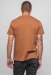 T-shirt short marron à manches courtes en coton doux, avec un col rond et une coupe décontractée, porté avec un jean noir. Tatouage visible sur le bras.