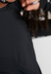 Černé sportovní oblečení s jemnými logy, hladkou texturou a přiléhavým střihem, s rozostřeným pozadím a viditelnými detaily ruky.