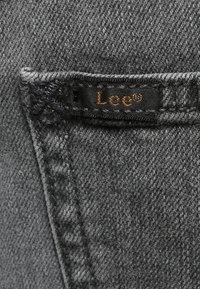 Tessuto in denim grigio scuro con una cucitura visibile e un'etichetta nera cucita con il nome del marchio "Lee" in oro.