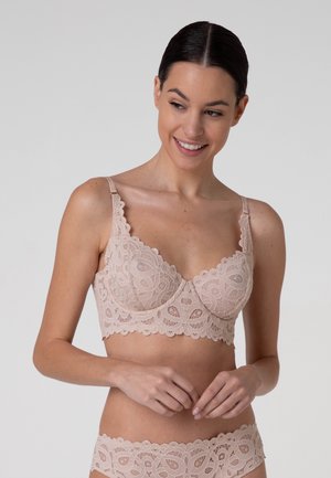 Donna che indossa reggiseno e mutandine in pizzo beige abbinati, sorridente, con capelli scuri raccolti all'indietro su uno sfondo grigio uniforme.