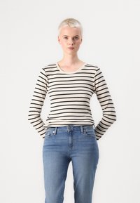 Persona con cabello rubio corto que viste una camiseta de manga larga a rayas beige y negra y jeans azules, contra un fondo blanco liso.