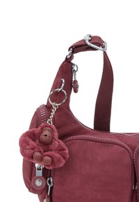 Lille maroon skuldertaske med justerbar rem, lynlås lommer og en blød gorilla nøglering hængende på frontlynlåsen.