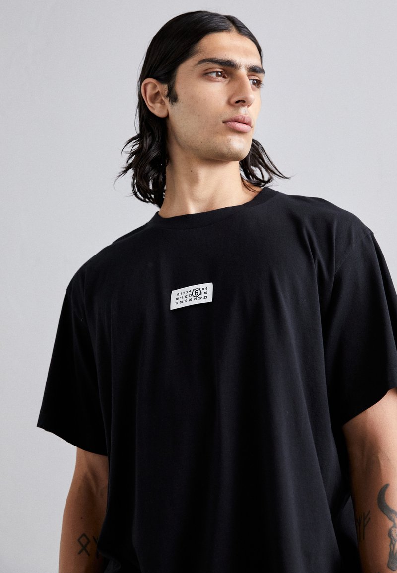 T-shirt oversize in cotone nero con etichetta bianca rettangolare stampata sul petto con numeri. Tessuto morbido, maniche larghe.