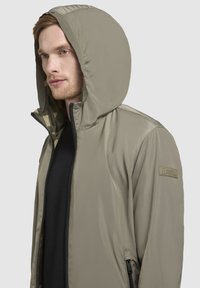 Kapuzejacke in hellolivgrün, aus glattem, leichtem Material mit Reißverschluss und seitlichen Taschen; weist ein Logopatch am Arm auf.