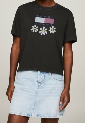 Camiseta negra corta con el texto "TOMMY JEANS" y un gráfico floral que presenta pétalos blancos y centros amarillos, combinada con una falda de mezclilla clara.