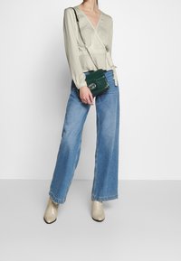 Maglione grigio chiaro con maniche lunghe, abbinato a jeans blu in denim a gamba larga. Tiene in mano una borsa verde in eco-pelle effetto coccodrillo e indossa stivaletti beige.