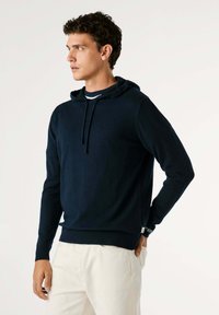 Modra navy kapuca iz mehkega materiala z sprednjim žepom, kapuco z vrvico in rebrastimi manšeti. Nosi se z svetlimi hlačami, preprost dizajn.