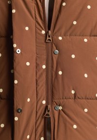Braune Steppjacke mit beigen Polka-Dots, mit zentralem Reißverschluss und Druckknöpfen entlang der Vorderseite.