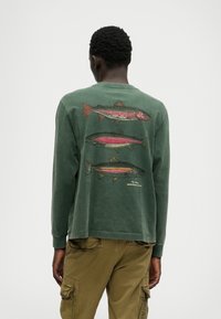 Vihreä pitkähihaisessa paidassa on selkäpuolella kolme yksityiskohtaista kalan kuvastoa pinkissä, mustassa ja vihreässä värissä, ja se on valmistettu pehmeästä kankaasta.