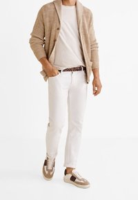 Cardigan en tricot beige, t-shirt clair, pantalon blanc slim et baskets avec accents marron. Le cardigan comporte un col châle et des boutons.