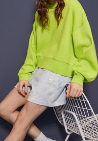 Neonově zelená oversize mikina s kulatým výstřihem, doplněná světle modrými kraťasy s batikovým vzorem a bílým logem. Modelka sedí na drátěné židli.