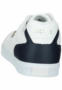 Sneaker alb cu toc bleumarin, partea superioară din piele și logo-ul aurit "LACOSTE". Dispune de o talpă din cauciuc și emblemă cu crocodilul semnătură pe lateral.