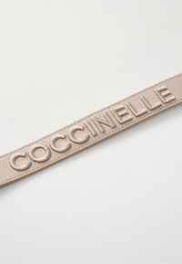 Bež kožni remen utisnut s "COCCINELLE", s teksturiranom površinom i glatkim završetkom, postavljen pod kutom horizontalno preko jednostavne pozadine.