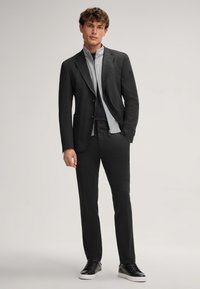 Blazer nero su misura con due tasche frontali, camicia grigia con zip sotto, pantaloni neri abbinati e sneakers nere con suola bianca.