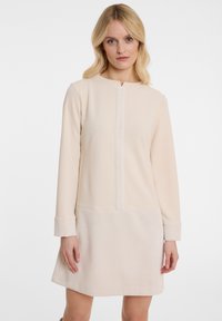 Beiges Kleid mit langen Ärmeln, das über ein strukturiertes unteres Panel und einen glatten oberen Abschnitt verfügt. Akzente bilden eine dezente zentrale Nahtdetail.