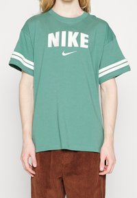 Grön bomull t-shirt med vit "NIKE" logotyp och swoosh, med vita ränder på ärmarna. Rund hals, korta ärmar, avslappnad passform.
