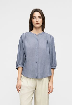 Vrouw met een blauw blouse met krijtstreepjes en knopen, driekwart pofmouwen en beige broek met hoge taille, staande tegen een effen achtergrond.
