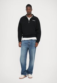 Zwarte fleece zip-up sweatshirt met geribbelde manchetten, gecombineerd met lichtblauwe denimjeans en witte sneakers, staande tegen een effen achtergrond.