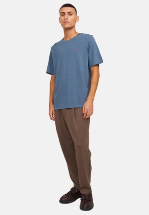 Homme debout portant un T-shirt bleu à manches courtes, un pantalon ample marron et des chaussures marron foncé sur un fond blanc uni.