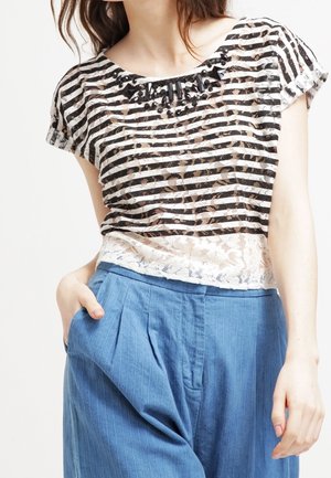 Vrouw die een doorschijnende zwart-wit gestreepte kanten top met korte mouwen draagt en blauwe, hoge taille, geplooide broek, met één hand in de zak.