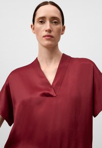 Blusa di satin borgogna con maniche corte e scollatura a V, caratterizzata da una texture liscia e un design minimalista.