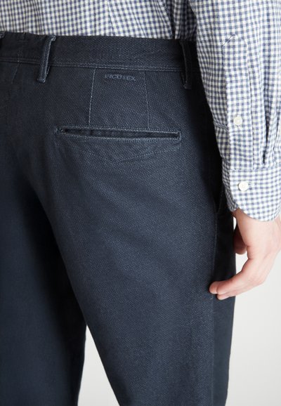 Pantalones navy hechos de un tejido texturado, con un bolsillo trasero y un sutil logo de la marca. Llevados con una camisa de cuadros azul y blanca.