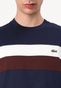 Μπλε ναυτικό μπλουζάκι με οριζόντιες ρίγες σε λευκό και μπορντό, με κεντητό λογότυπο κροκόδειλου σε πράσινο χρώμα στο επάνω αριστερό μέρος του στήθους.