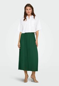 Camicia bianca corta con bottoni e maniche corte, abbinata a una lunga gonna plissettata verde e sandali metallici con cinturino.