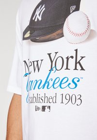 Camiseta blanca con un gráfico de una gorra de béisbol azul marino y una pelota de béisbol. El texto dice "New York Yankees Establecidos en 1903" en negro y azul.