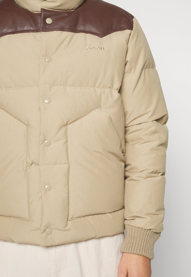 Giubbotto imbottito beige con accenti in pelle marrone sulle spalle, due grandi tasche frontali e chiusura a scatto. Polsini a costine di un colore più chiaro.