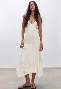 Abito lungo bianco in pizzo con scollatura a V, caratterizzato da strisce verticali testurizzate e orlo fluente. Indossato con sandali neri flats.