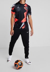 Man som står i en svart och röd Paris Saint-Germain fotbollströja med svarta Nike-byxor och vita fotbollsskor, med en svart och orange fotboll på golvet.