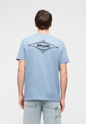 Mężczyzna ubrany w jasnoniebieską koszulkę Ripcurl i jasne dżinsy, widoczny od tyłu na jednolitym tle.