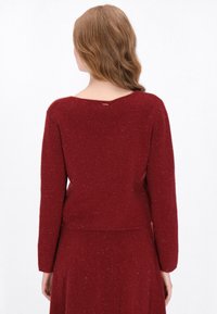 Rode gebreide trui met een ronde achterkant van de halslijn, lange mouwen en een iets verkorte zoom. Zachte textuur met subtiele glanselementen.