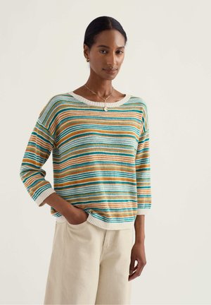 Frau mit dunklen Haaren, die goldene Creolen, eine goldene Halskette, einen gestreiften Pullover in Blau, Grün und Orange sowie hellbeige Hosen trägt, mit einer Hand in der Tasche.