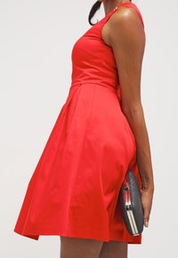Rotes ärmelloses Kleid mit tailliertem Oberteil und ausgestellt Rock; glattes Satinmaterial. Schwarze runde Clutch in der rechten Hand gehalten.