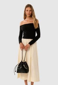 Frau in einem schwarzen schulterfreien Oberteil und einem cremefarbenen Midikleid, die eine schwarze Zugband-Eimer-Tasche mit dem Schriftzug "FOREVER NEW" hält.