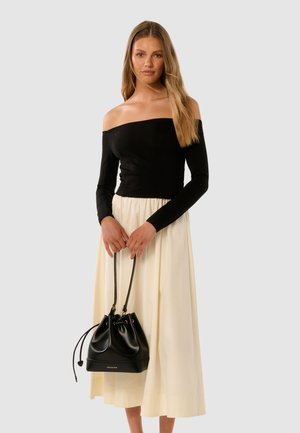 Kvinne med svart off-shoulder topp og kremfarget midi-skjørt, som holder en svart snøre-buksepose med "FOREVER NEW" trykt på.
